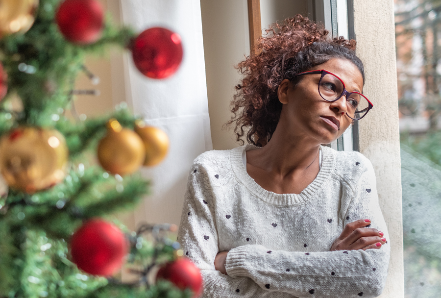 Woman with sad face looking out window and Christmas tree in foreground